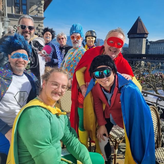Sujet 2023: Helden — Musiker in bunten Superheldenkostümen mit Umhängen und Masken, im Hintergrund der Wasserturm