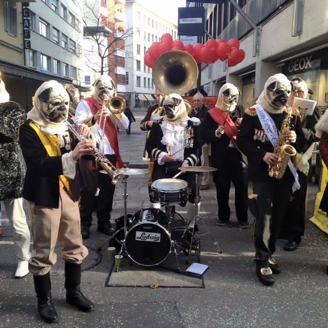 Sujet 2016: Möps — Auf den Hund gekommen — die Band mit Mopsmasken in der Gasse, dahinter rote Herzballone